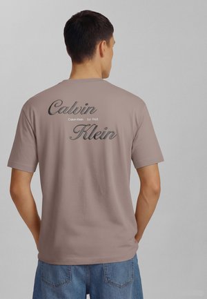 Man draagt taupekleurige Calvin Klein T-shirt met groot zwart schriftlogo op de achterkant, gecombineerd met blauwe denimshorts, staand tegen een eenvoudige grijze muur.