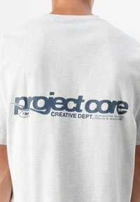 Un primer plano de la espalda de una persona que lleva una camiseta gris claro con "project core" y "CREATIVE DEPT." impresos en azul oscuro.
