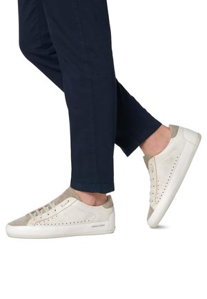 Weiße Wildledersneaker mit beigen Akzenten und durchbrochener Detailierung. Flache Gummisohle. Getragen mit navyblauen Hosen, was einen lässigen Stil zeigt.
