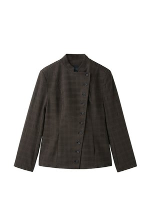 Femme portant des lunettes de soleil surdimensionnées, une veste marron boutonnée, un jean à jambes larges, des chaussures blanches pointues, tenant un sac à main vert sur un fond rose uni.