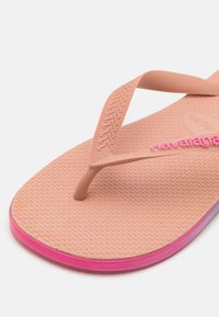 Vaaleanpunaiset ja beige kumiset sandaalit, joissa on tekstuuroitu pohja, kapeat hihnat ja logot yksityiskohta. Pohjassa on gradienttiväri.