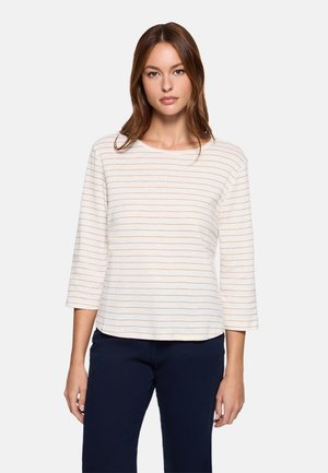 Mujer con una blusa blanca de manga larga con finas rayas horizontales beige y pantalones azul marino oscuro, de pie contra un fondo claro y liso.