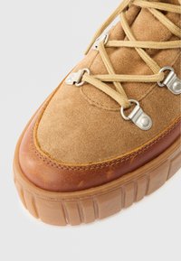 Barna velúr sneaker, világosbarna bőr kiegészítőkkel, fémből készült fűzőlyukakkal és kerek bézs fűzőkkel. Vastag, texturált gumitalp barázdákkal.