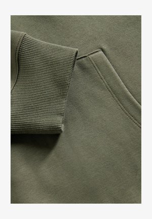 Detailní záběr na tkaninu olivově zelené mikiny, ukazující žebrovaný rukáv a švy na přední kapse.