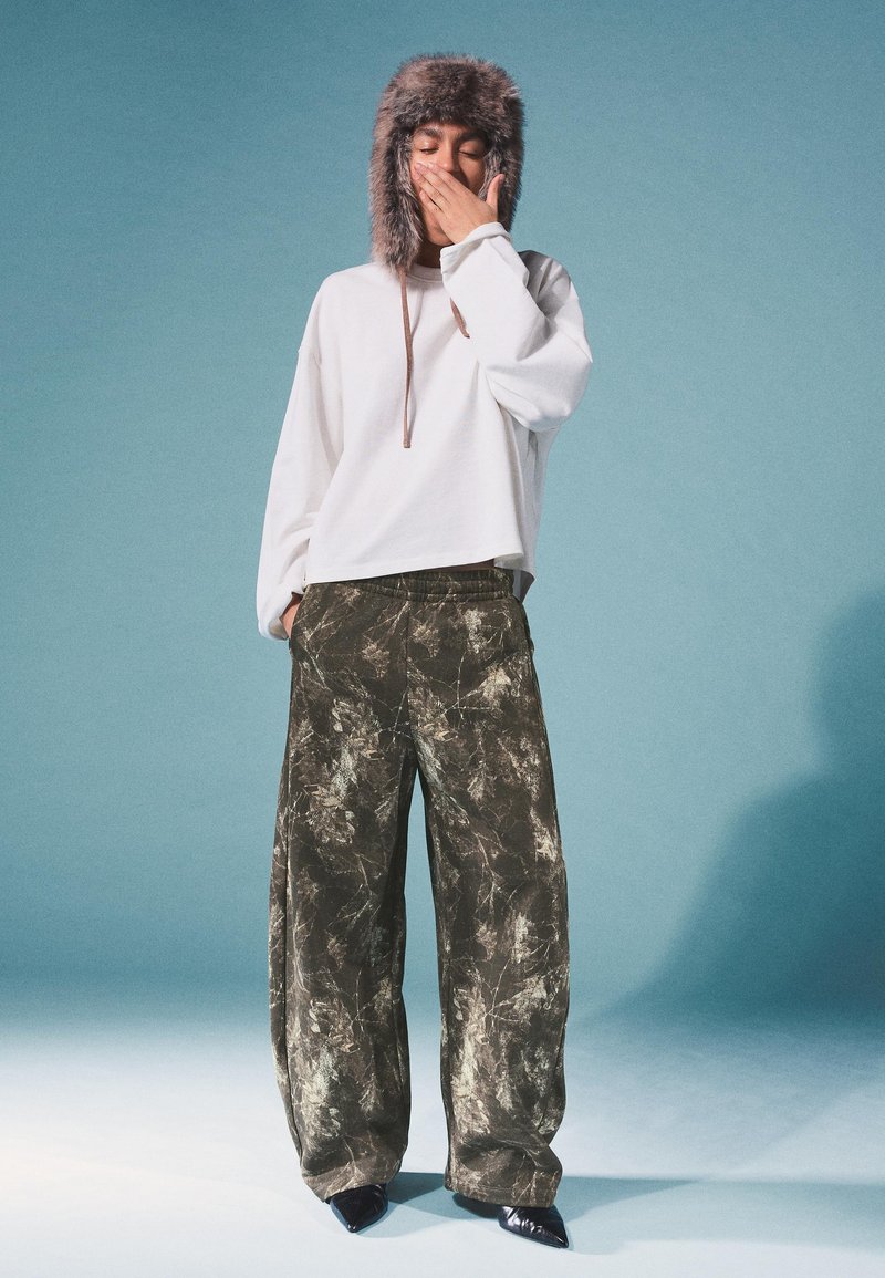 Biały krótki sweter, zielono-brązowe spodnie w szerokie nogawki w marmurkowy wzór oraz puszysta, brązowa czapka z uszami. Tło jest jasnoniebieskie.