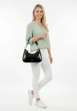 Blonde Frau, die eine hellgrüne Bluse, weiße Hose und weiße Sneakers trägt, eine glänzende schwarze Handtasche über der Schulter hält und lächelt.