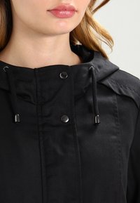 Svart huva jacka med dragkedja framtill, justerbara dragsko och tryckknappdetaljer på kragen. Slät tygstruktur.