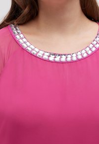 Blouse rose avec un col arrondi orné d'une rangée de pierres transparentes. Le tissu est lisse et semi-transparent au niveau des épaules.