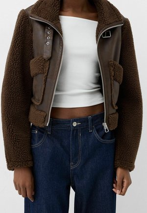 Person iført en brun shearlingjakke med lynlås og spænde detaljer over en hvid crop top og blå højttaljede jeans.