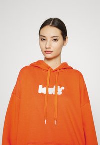 Joven con cabello oscuro recogido, vistiendo una sudadera naranja de Levi's con cordones, sobre un fondo claro y simple.