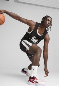 Débardeur de sport noir avec des accents blancs, shorts à motif, et sneakers rouges, noirs et blancs. Basketball dans la main droite, en pose d'action.