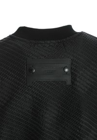Chaqueta negra texturizada con un patrón geométrico, cuello acanalado y una etiqueta rectangular de cuero con botones de presión en la parte posterior.