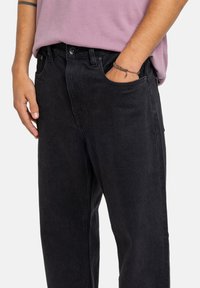 Jeans in denim neri con vestibilità comoda, dotati di chiusura con bottone, passanti per la cintura e tasche laterali. Indossati con una camicia di colore lilla chiaro.