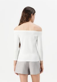 Top bianco a spalle scoperte e maniche lunghe, con una siluette aderente e una texture liscia; abbinato a pantaloni corti grigi chiari.