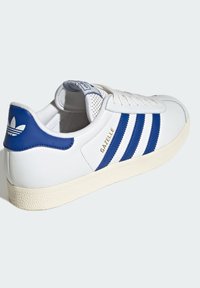 Bijela tenisica s plavim akcentima i tri pruge. Ima teksturiranu gumenu podlogu i podstavljeni jezik s mrežastom oblogom. "Gazelle" u zlatnoj boji.