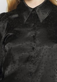 Musta satiini pusero, jossa on kukkakuvio, terävä kaulus ja kuusi mustaa nappia edessä, sileä pinta.