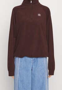 Brun fleecepullover med hög krage, halvdragkedja fram och Adidas-logga på bröstet. Matchas med ljusblå vida jeans.