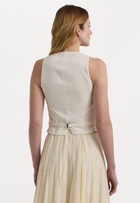 Top sem mangas bege com um design ajustado, a textura do tecido é suave, apresentando um detalhe de fecho nas costas. Usado com uma saia clara e plissada.
