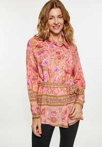 Blusa floreale rosa con colletto, maniche lunghe e vita legata. Presenta motivi intricati e dettagli dorati. Pantaloni neri.