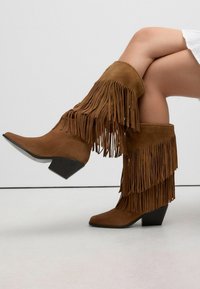 Met gekruiste benen en mid-calf bruine suède laarzen met gelaagde franjes en lage blokhakken tegen een effen grijze achtergrond.