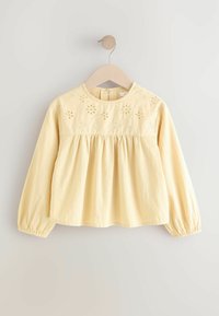 Gul bomuldsbluse med lange ærmer, samlet talje og blomsterbroderi på skuldrene. Knappelukning bagpå og elastiske manchetter.