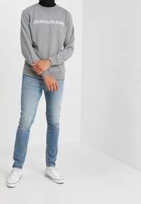 Grå sweatshirt med "CALVIN KLEIN JEANS" tryckt i vitt, i kombination med ljusblå jeans och vita sneakers. Fitted design med rund hals.