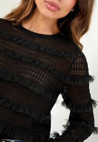 Schwarzes Langarmoberteil mit strukturierten Streifen und transparenten Rüschendetails, das ein gestricktes Muster und eine figurnahe Passform zeigt.