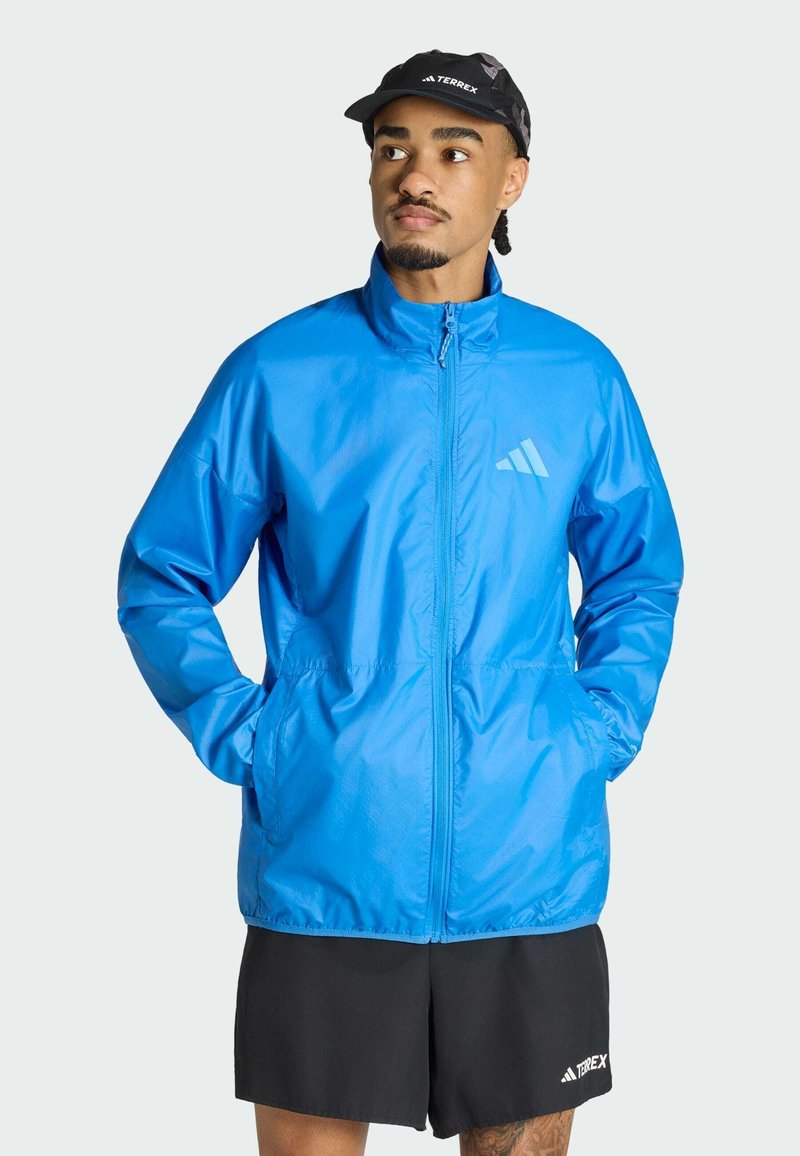Homme portant une veste zippée bleu vif, un short noir et une casquette noire, debout les mains dans les poches, regardant sur le côté.