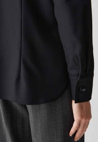 Camicia nera con scollatura e polsini strutturati, con una singola chiusura a bottone. È abbinata a pantaloni grigi a righe.