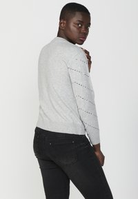 Maglione grigio chiaro con motivo testurizzato sulle maniche, scollatura rotonda e orlo a coste; abbinato a jeans neri. Messa in risalto la varietà dei tessuti.