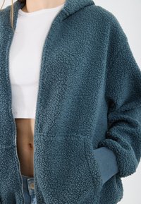Teal fleece zip-up hoodie met een gestructureerd oppervlak, voorzien van een capuchon, voorzakken en geribbelde manchetten. Eronder een witte getailleerde shirt.