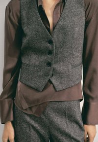 Chaleco de lana gris con botones negros superpuesto a una camisa de satén marrón, que presenta mangas largas y una textura suave. Se pueden ver pantalones a juego.