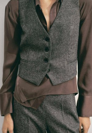 Chaleco de lana gris con botones negros superpuesto a una camisa de satén marrón, que presenta mangas largas y una textura suave. Se pueden ver pantalones a juego.