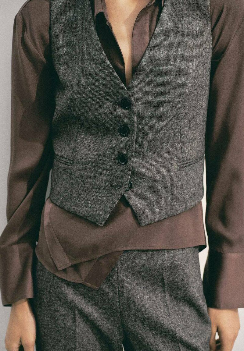 Chaleco de lana gris con botones negros superpuesto a una camisa de satén marrón, que presenta mangas largas y una textura suave. Se pueden ver pantalones a juego.