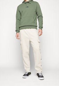 Felpa verde con logo, pantaloni cargo crema con tasche laterali e bottoni a pressione, sneakers nere con suole bianche e accenti a fantasia.