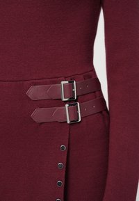 Maroona pletena haljina s dva usklađena remena od umjetne kože sa srebrnim kopčama u struku i dekorativnim dugmadi na bočnoj strani.