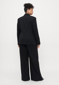 Veste de costume noire ajustée avec un col aiguisé et des épaules structurées, associée à un pantalon à jambes larges ; tissu lisse et lignes épurées.