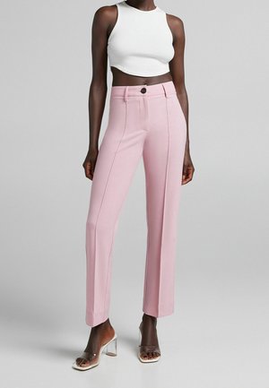 Pantalons ajustés rose clair à taille haute, jambes droites et détails de couture frontaux. Assortis à un crop top blanc et des sandales à talons transparents.