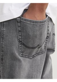 Grå denimjeans med et nærbillede af baglommen. Har diskrete syninger og en tekstureret overflade. Skjortens kant er delvist synlig ovenover.