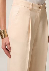 Helle beigen Hosen aus glattem Stoff, mit seitlichen Falten, einem Knopfverschluss und einem taillierten Schnitt. Ein goldenes metallisches Armband ist sichtbar.