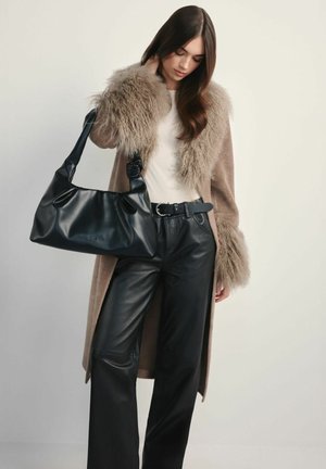 Kobieta ubrana w beżowy płaszcz z owczej skóry z futrzanymi mankietami, białą bluzkę, czarne skórzane spodnie, czarny pasek, trzyma dużą czarną skórzaną torebkę.