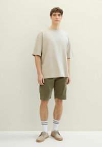 Beżowa, oversize'owa koszulka, oliwkowe szorty cargo, białe skarpetki w czarne paski i beżowe sneakersy, na neutralnym tle.