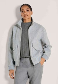 Frau, die eine hellgraue Jacke mit Reißverschluss und großen Taschen, einen dunkelgrauen strukturierten Pullover und graue Hosen trägt. Sie steht vor einem schlichten Hintergrund.