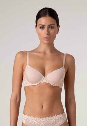 Donna in piedi e rivolta in avanti, indossa un completo coordinato di reggiseno e slip color carne con dettagli in pizzo, sfondo neutro.