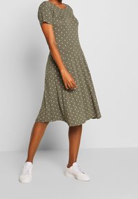 Oliwkowa sukienka z krótkim rękawem w białe kropki, z rozszerzanym dołem, wykonana z miękkiego materiału. Połączona z białymi sneakerami.