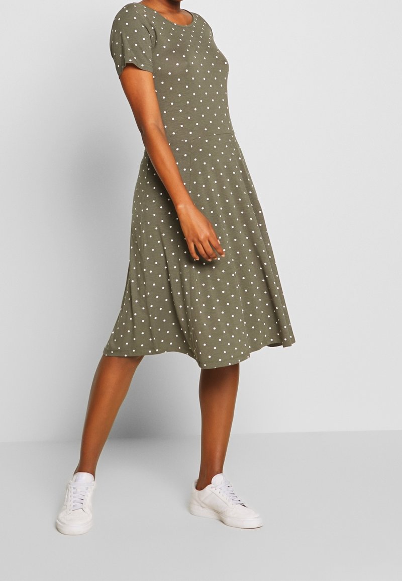 Oliwkowa sukienka z krótkim rękawem w białe kropki, z rozszerzanym dołem, wykonana z miękkiego materiału. Połączona z białymi sneakerami.