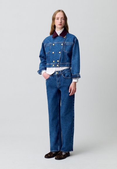 Giacca di jeans blu con bottoni dorati, sovrapposta a una camicia bianca con colletto. Abbinata a pantaloni larghi in denim blu e mocassini marroni a fantasia.