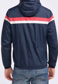 Námořnická větrovka s kapucí, zdobená vodorovnými červenými a bílými pruhy přes ramena, s elastickými manžetami a staženým lemem.