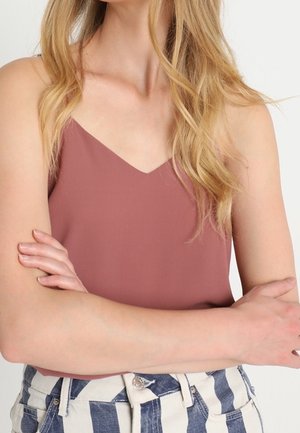 Kobieta z założonymi skrzyżowanymi ramionami, ubrana w fioletowy top bez rękawów oraz wysokie, niebiesko-białe spodnie w paski.