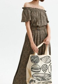 Brun mönstrad axelbandslös klänning med volangärmar och åtdragen midja. Modellen håller en krämfärgad tote-väska med svarta botaniska illustrationer.